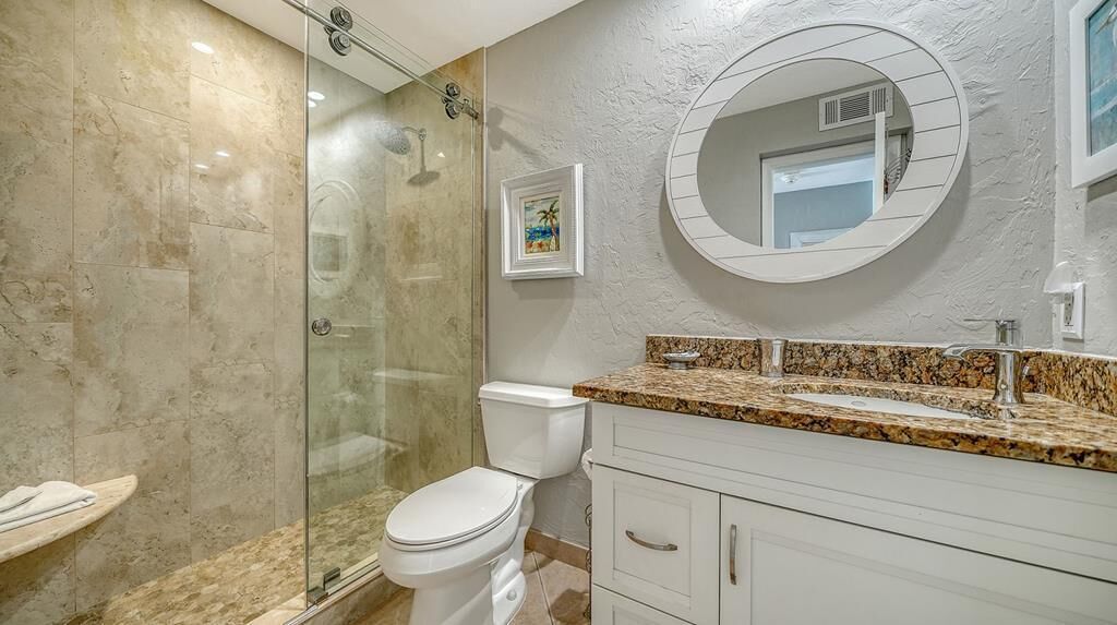 A bathroom with a glass-enclosed shower, a toilet, a sink with a granite countertop, and a large mirror.