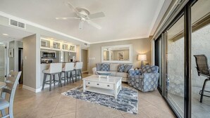 A modern living room with a white coffee table, a sofa, and a TV. There is a kitchen area with a microwave and a dining table.