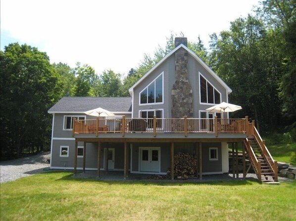 A two-story house with a stone chimney, a deck with outdoor furniture, and a staircase leading to the upper level.
