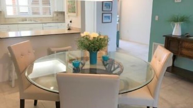 Un espace repas moderne avec une table en verre et des chaises blanches, un vase rempli de fleurs et, au fond, une cuisine.