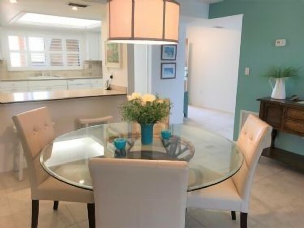 Un espace repas moderne avec une table en verre et des chaises blanches, un vase rempli de fleurs et, au fond, une cuisine.