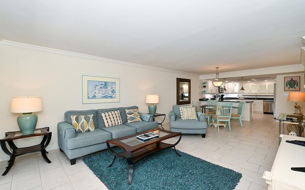 A living room with a sofa, coffee table, and lamps. There is a dining area with chairs and a table. The kitchen is visible in the background.