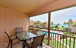 A balcony with a glass table and chairs, overlooking a pool and a landscaped area with palm trees.