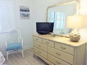 A room with a dresser, a television, a chair, a lamp, and a mirror.