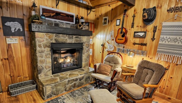 Ein gemütliches Blockhaus-Interieur mit Kamin aus Stein, Holzwänden, einem Schaukelstuhl, einer Bank und einer Gitarre an der Wand.