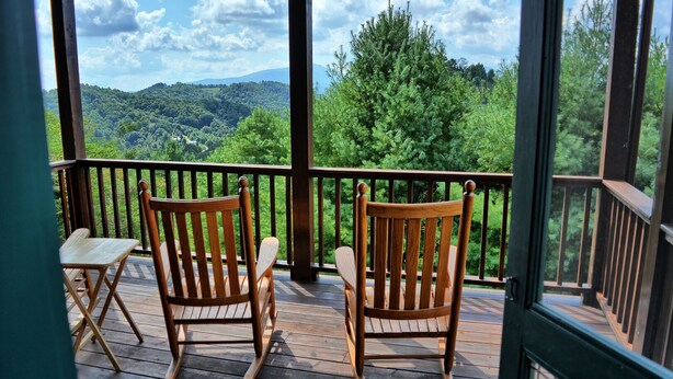 Um deck de madeira com duas cadeiras de balanço e uma mesa, com vista para uma paisagem arborizada.
