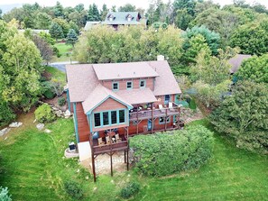 A large two-story house with a balcony and a deck, surrounded by lush greenery.