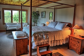Ein Himmelbett mit floralem Überwurf, ein Nachttisch mit Lampe, eine Holzbank und Blick ins Freie durch große Fenster.