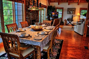 A dining area with a table set for six, surrounded by wooden chairs, a stone wall, and a television in the background.