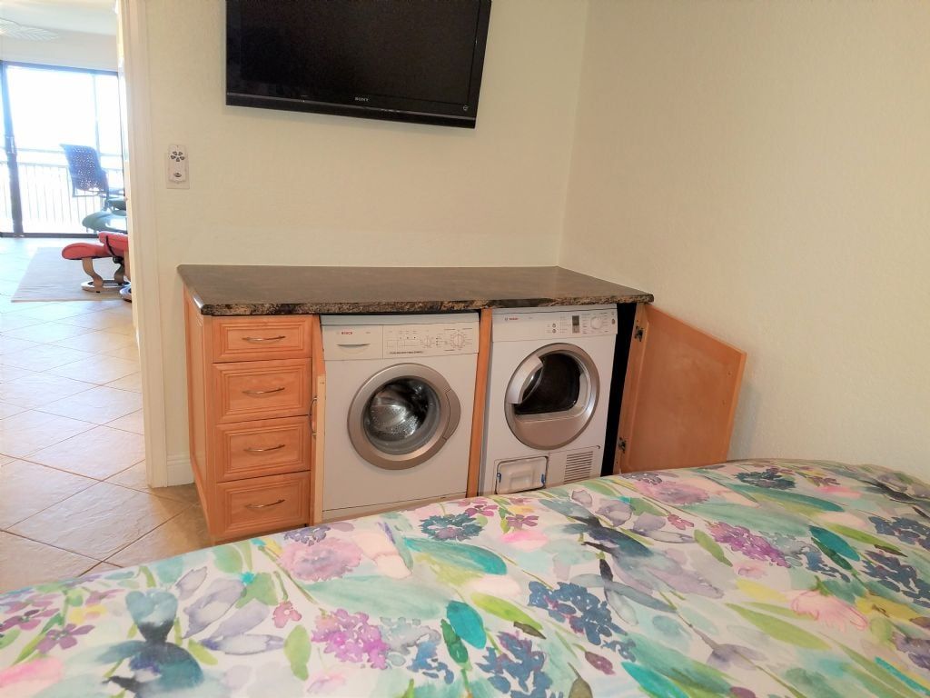 Une chambre à coucher avec un lit, un lave-linge, un sèche-linge, une armoire en bois et un téléviseur fixé au mur.