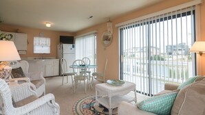 A cozy living room with wicker furniture, a dining table, and a view of the outside through large windows. - 385 Cute 3 Bedroom Cottage in South Nags Head (Nags Head)