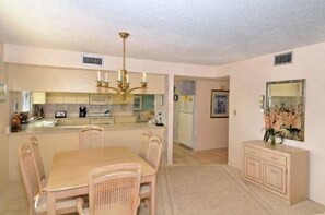 A dining area with a table and chairs, a kitchen in the background, and a hallway leading to another room.