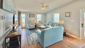 A living room with a blue sofa, two armchairs, a coffee table, a ceiling fan, and a view of the ocean through a sliding door. - Riki Tiki | Oceanfront Home in Surf City (Surf City)