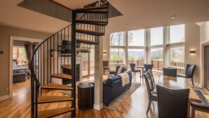 A spacious living area with a wooden staircase, a fireplace, a dining area with a table and chairs, and large windows offering a view of the outdoors.