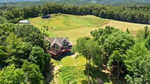 A large house with a prominent front porch and multiple windows, surrounded by a lush green landscape and forested hills in the background.