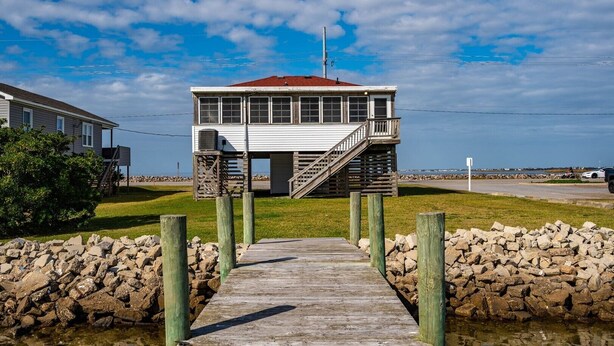 A wooden pier leading to a white building with a red roof, situated on a grassy area with a stone wall on one side. - 275 Unique Water-Front Home w/ Private Pier (Nags Head)
