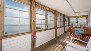 A covered porch with large windows, wooden flooring, and a dining table with chairs. - 275 Unique Water-Front Home w/ Private Pier (Nags Head)
