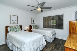 Une chambre avec deux lits, un ventilateur de plafond, une fenêtre avec des stores et une commode en bois.