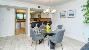 A dining area with a glass table surrounded by grey chairs, a modern chandelier, and a view of the kitchen in the background. - Chinaberry 932- 2 bedroom 2 full bath unit on 3rd floor overlooking bay (Siesta Key)