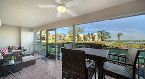 Ein sonnendurchfluteter Balkon mit Korbmöbeln, einem Deckenventilator und Blick auf eine angelegte Grünfläche mit Palmen und einem Gewässer.