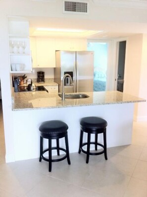 A modern kitchen with a stainless steel refrigerator, a built-in sink, and two black stools. - Firethorn 110- Absolutely gorgeous, beachfront 2BR/2BA condo (Siesta Key)