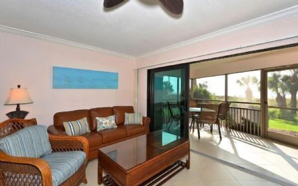 A living room with a sofa, armchair, and coffee table, a painting on the wall, and a balcony with outdoor furniture.