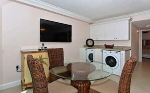 A compact living space with a glass-top dining table, wicker chairs, a television, a clock, and a washing machine. - Firethorn 110- Absolutely gorgeous, beachfront 2BR/2BA condo (Siesta Key)