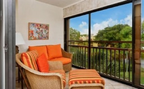A balcony with a wicker sofa and ottoman, a painting on the wall, and a view of greenery.