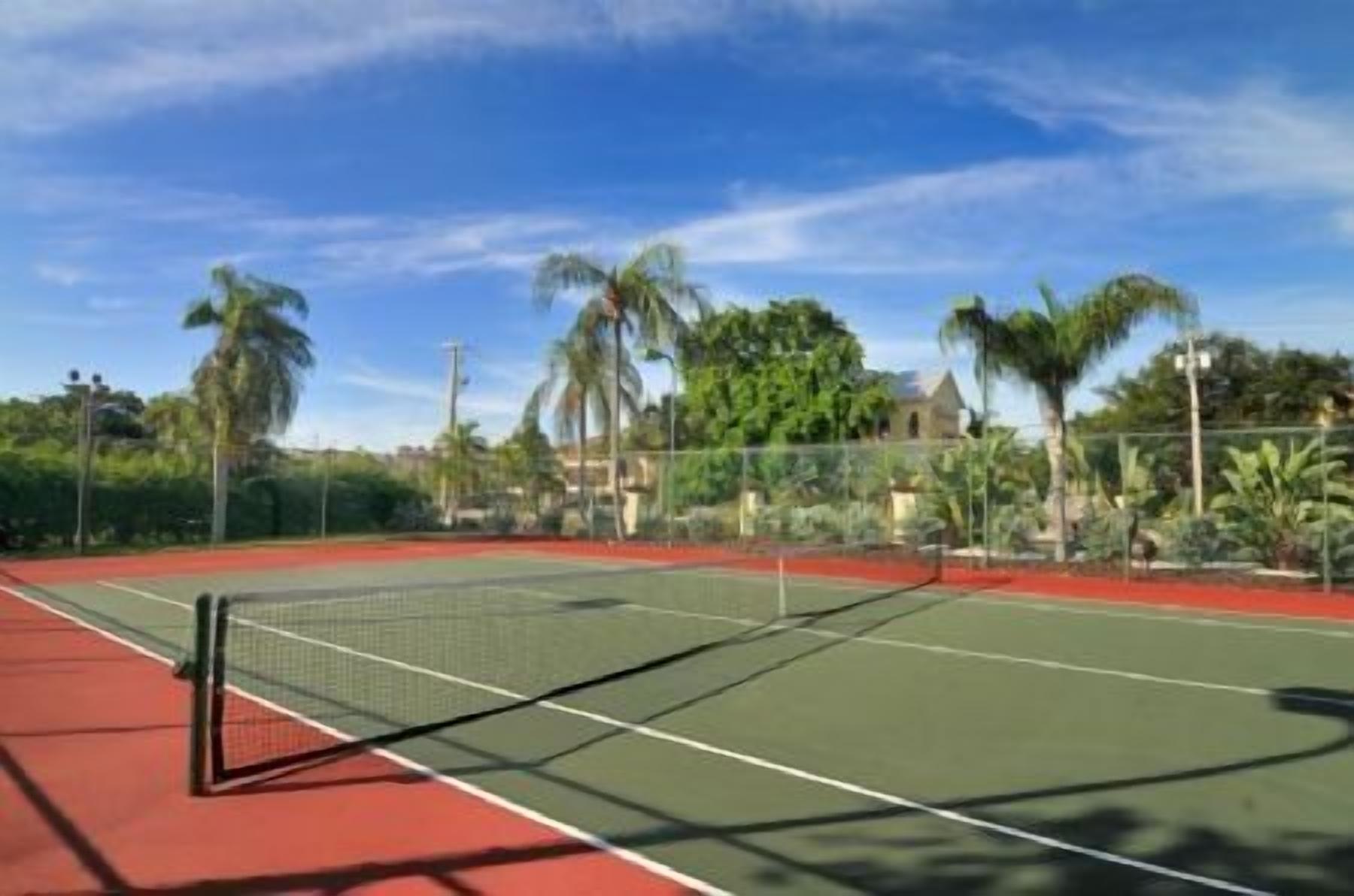 An outdoor tennis court with a net, surrounded by greenery and a clear sky.