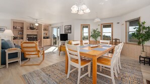 A spacious living area with a wooden dining table, white chairs, a sofa, and a TV. There is a kitchen area with wooden cabinets and a fireplace. Large windows provide natural light and views of the outdoors.