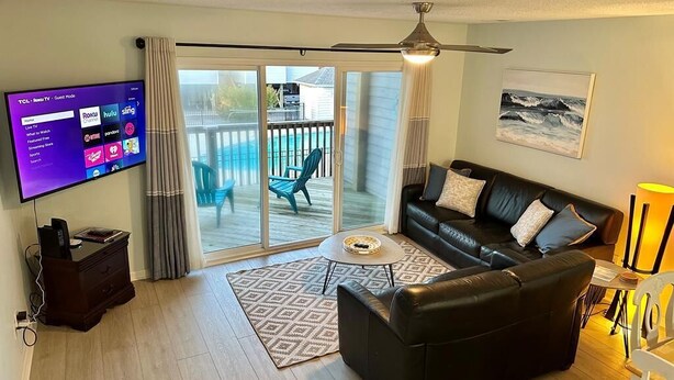 A modern living room with a flat-screen TV, a black leather sofa, a coffee table, and a sliding glass door leading to a balcony. - Turtle Cove 133 | Surf City, NC | Community Pool & Beach Access (Surf City)