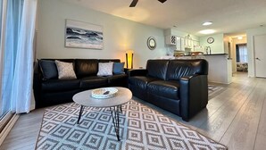 A modern living room with a black leather sofa, a round coffee table, and a patterned rug. A ceiling fan is visible, and the room has light-colored walls and wooden flooring. - Turtle Cove 133 | Surf City, NC | Community Pool & Beach Access (Surf City)