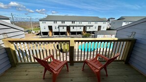 A wooden deck with two red chairs, a pool in the background, and a row of white buildings. - Turtle Cove 133 | Surf City, NC | Community Pool & Beach Access (Surf City)