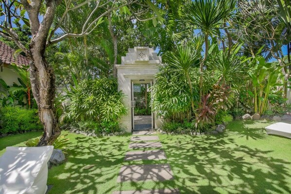 A garden pathway leading to a white structure with a door, surrounded by lush greenery and palm trees.