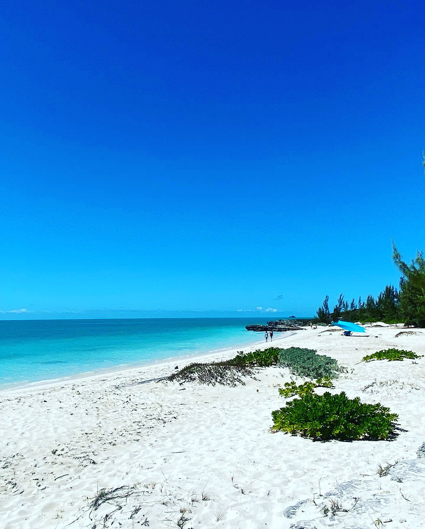 Uma praia com areia branca, águas azul-claras e algumas pessoas caminhando.