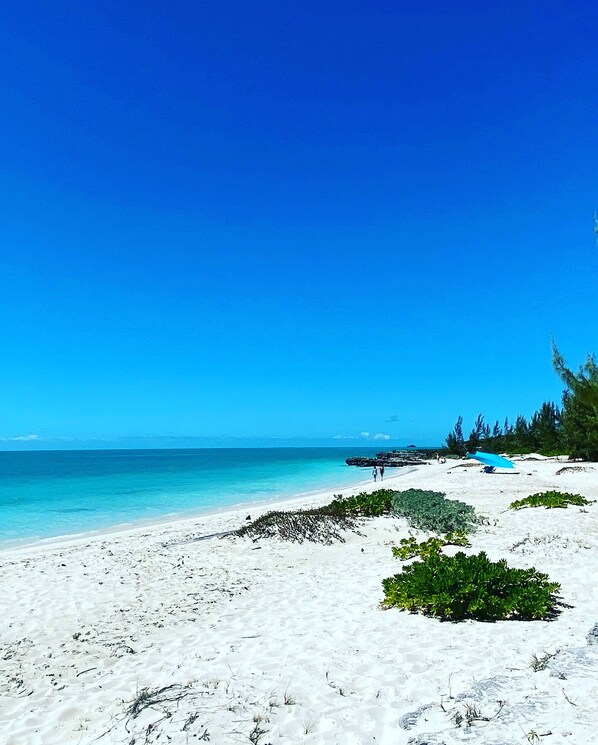 A beach with white sand, clear blue water, and a few people walking.