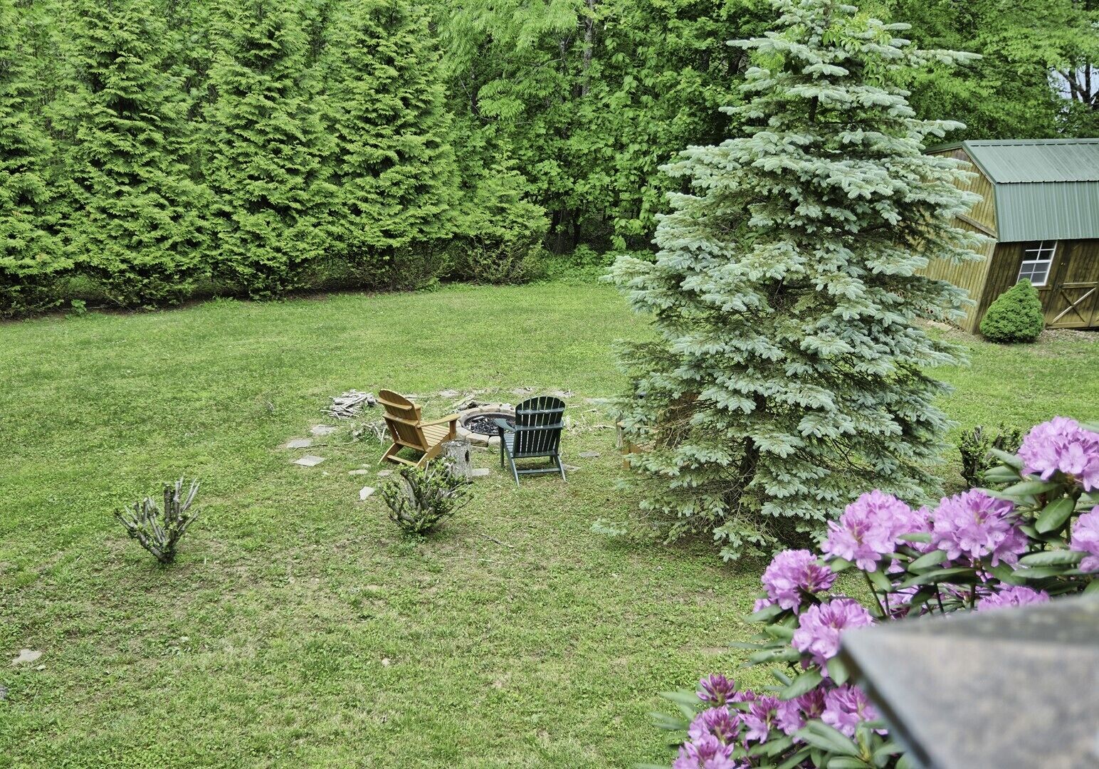 A grassy backyard with a wooden chair, a metal chair, and a small fire pit.