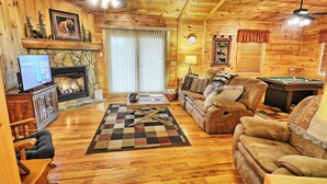 A cozy cabin interior with a fireplace, wooden floors, a sofa, a pool table, and ceiling fans.