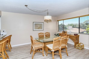 A dining area with a glass table surrounded by wicker chairs, a chandelier, a framed picture on the wall, and a window with blinds. - Doveplum 621- Newly remodeled and refurnished 2BR/2.5BA townhome (Siesta Key)