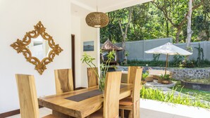 Uma mesa de jantar de madeira com cadeiras, um espelho decorativo e um candeeiro pendente num quarto com vista para um jardim e um pequeno pavilhão.