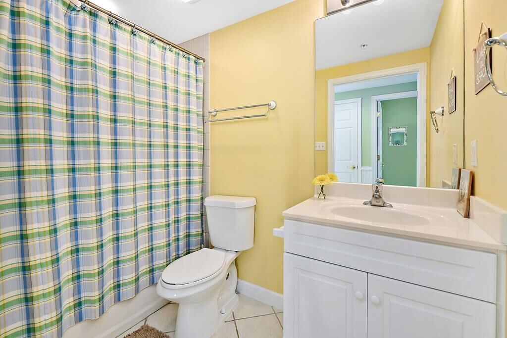 Une salle de bain avec un rideau de douche à carreaux, des toilettes blanches, un lavabo blanc avec un miroir et un mur jaune.