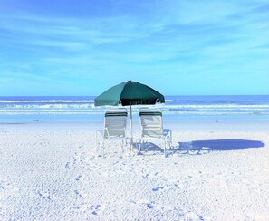 A beach with two chairs under a green umbrella, facing the ocean.