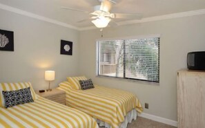 Une chambre avec deux lits, un ventilateur de plafond, une fenêtre avec des stores, une lampe de chevet et un téléviseur posé sur un meuble.