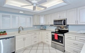 A modern kitchen with white cabinets, a stainless steel dishwasher, a microwave, an oven, and a ceiling fan. - Chinaberry 456-Amazing sunset views every night from this 5th floor 2BR/2BA condo (Siesta Key)