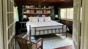 Een slaapkamer met een houten plafond, een metalen bedframe, een nachtkastje met een lamp, een stoel, een boekenplank en een raam met gordijnen.