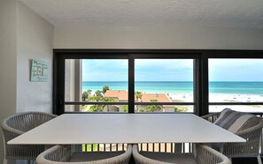 A room with a large window overlooking a beach, a white table, and wicker chairs.