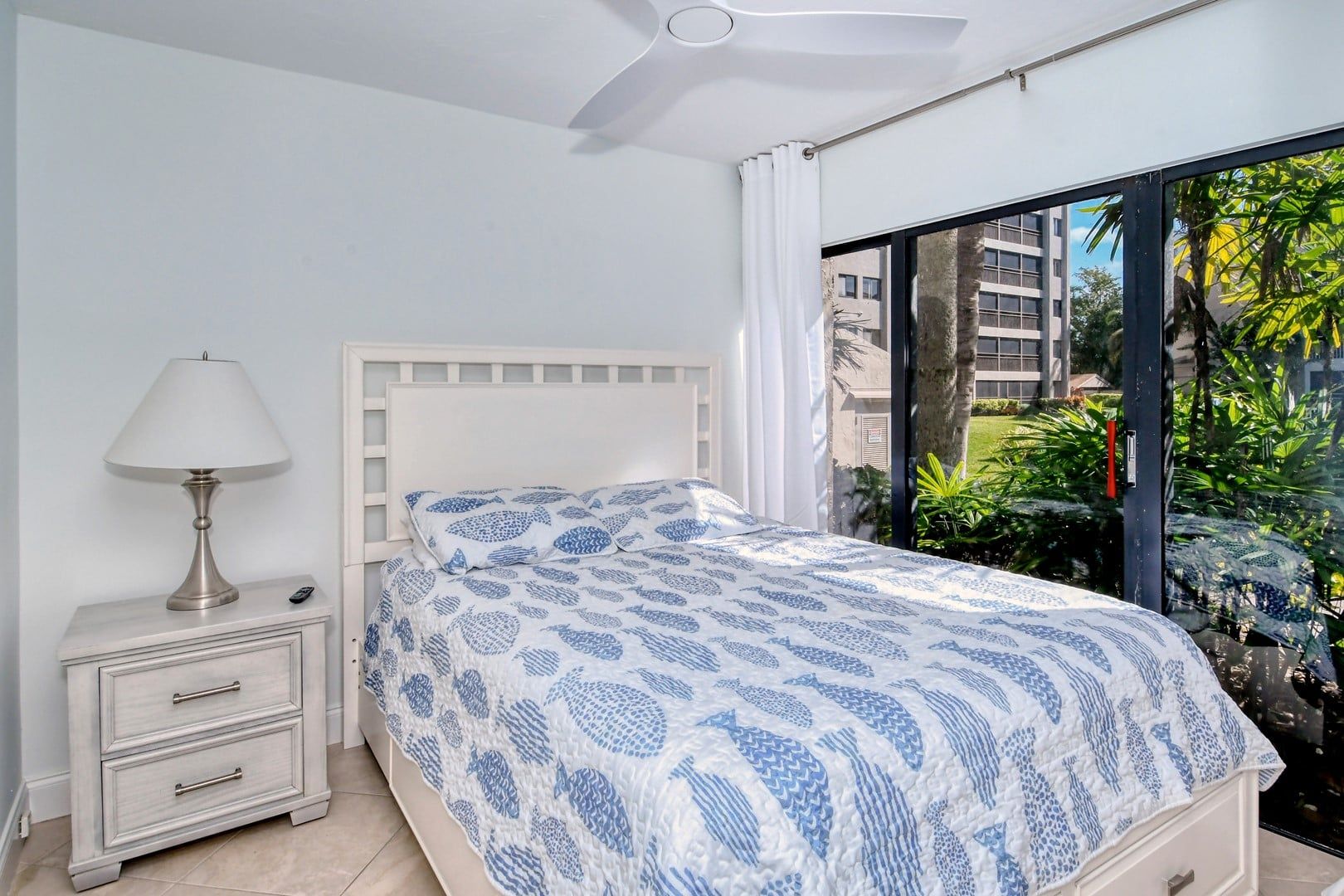 A bedroom with a bed, a bedside table with a lamp, and a view of greenery through a large window.