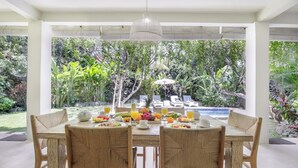 Un área de comedor al aire libre con una mesa dispuesta para dos personas, rodeada de abundante vegetación y una piscina al fondo.