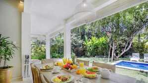Un comedor con una mesa lista para el desayuno, rodeado de exuberante vegetación y una piscina.