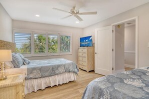 Ein Schlafzimmer mit zwei Betten, einem Deckenventilator, einem Fernseher und einem Fenster mit Blick auf Palmen.